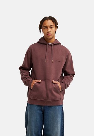 Junger Mann mit geflochtenen Haaren, der einen braunen Hoodie und blaue Jeans trägt, steht mit den Händen in der Hoodie-Tasche vor einem schlichten Hintergrund.