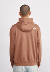 Sudadera con capucha marrón, con puños y dobladillo de canalé, textura suave, logo en la parte posterior y corte relajado. No hay detalles visibles en la parte delantera.