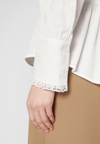 Main reposant à côté d'une jupe beige, portant un chemisier blanc à manches longues avec des poignet boutonnés et de la dentelle aux manches.