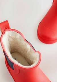 Czerwone gumowe botki z zaokrąglonym noskiem, z kremowym futerkiem w środku i granatowym akcentem z materiału. Zawierają pętelkę na pięcie dla łatwego zakładania.