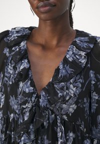 Femme portant un chemisier noir à imprimé floral bleu et blanc avec un col en V volanté, visible des épaules à la poitrine.