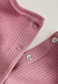 Rožnata rebrasta tkanina s srebrnimi pritiskači in okroglimi odprtinami za gumbe. Mehka tekstura, prikazuje bližnji posnetek robov oblačila.