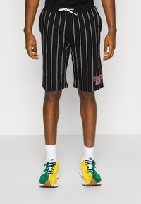Hombre vestido con pantalones cortos negros con rayas verticales blancas y el logo de "Tommy Jeans", calcetines blancos y zapatillas verde-amarillas, de pie sobre un suelo gris.