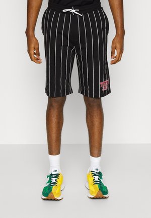 Hombre vestido con pantalones cortos negros con rayas verticales blancas y el logo de "Tommy Jeans", calcetines blancos y zapatillas verde-amarillas, de pie sobre un suelo gris.