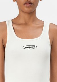 Λευκή ριγέ φανέλα με στρογγυλή λαιμόκοψη και λεπτές τιράντες, που διαθέτει το μαύρο λογότυπο "guess jeans" στην περιοχή του στήθους.