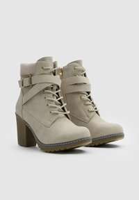 Botines beige de tacón alto, hechos de ante sintético, con suela texturizada, cordones, detalles de hebilla y un puño de punto.