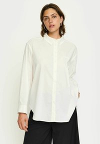 Cămașă albă cu mâneci lungi și închidere cu butoni, având o buzunare și un design subtil în formă de inimă. Material neted, croială lejeră.
