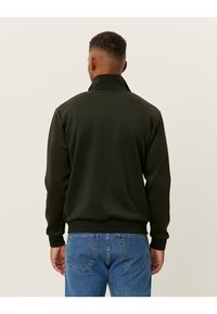 Mørkegrønn, zip-up jakke med høy krage og ribbestrikkede mansjetter. Glatt tekstur, tettsittende design, brukte med blå jeans. Enkel og moderne stil.