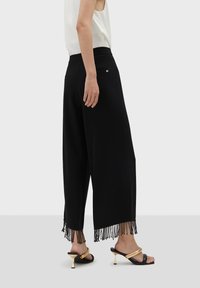 Svarta vida byxor gjorda av ett mjukt material, med franssömmar nedtill. En liten guldsymbol nära midjan. Matchas med svarta klacksandaler.