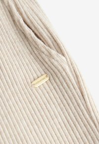 Ribbed knit-paita vaaleassa beige värissä, jossa on kultainen etiketti, jossa lukee "DOPPETIME". Pehmeä rakenne ja pitkänomainen muoto näkyvissä.