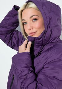Chaqueta inflable morada con capucha, que presenta una textura acolchada y cierre de cremallera. Cordón visible en el cuello y puños ajustados.