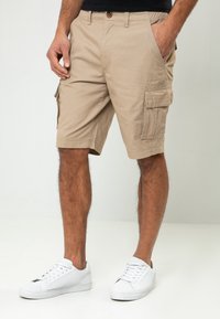 Shorts cargo beiges en coton, offrant une coupe décontractée, des poches latérales, une fermeture à bouton et un ourlet droit. Portés avec des baskets blanches.
