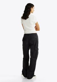 Pantalones cargo negros con un corte relajado, que presentan grandes bolsillos laterales, un cierre de botón y un detalle en la cinturilla. Combinados con una blusa blanca ajustada.