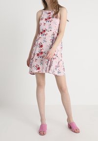 Blumenkleid in Rosa mit ärmellosem Design, verziert mit roten und blauen Blumen. Weicher Stoff mit einem ausgestellten Rock. Kombiniert mit rosa Absatzmules.