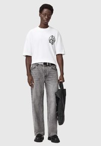 Valkoinen ylisuuri t-paita, jossa musta grafiikka rintamuksessa, yhdistettyinä löysiin harmaisiin farkkuihin ja mustiin kenkiin. Kannan olkalaukkua.