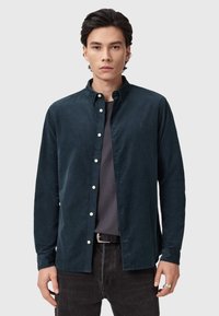Camicia in cotone a coste verde scuro con colletto button-down, bottoni frontali e un logo discreto. Indossata sopra una t-shirt grigia e jeans neri.