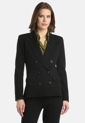 Vrouw in een zwarte dubbelgeknoopte blazer over een olijfgroene blouse met stippen, staand met haar handen ontspannen langs haar zij.