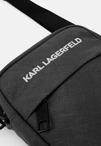 Szürke szövet keresztpántos táska fehér "KARL LAGERFELD" felirattal, cipzáros záródással és fekete állítható pánttal. Texturált felület.