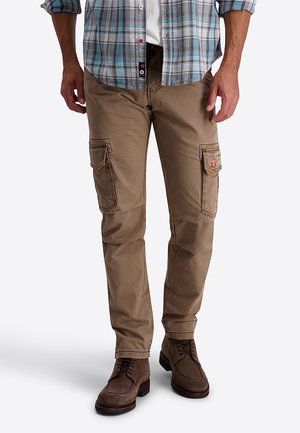 Mann trägt beige Cargohosen mit mehreren Taschen, braune Schnürstiefel, ein weißes Hemd und ein blau-graues kariertes Hemd mit Knöpfen.