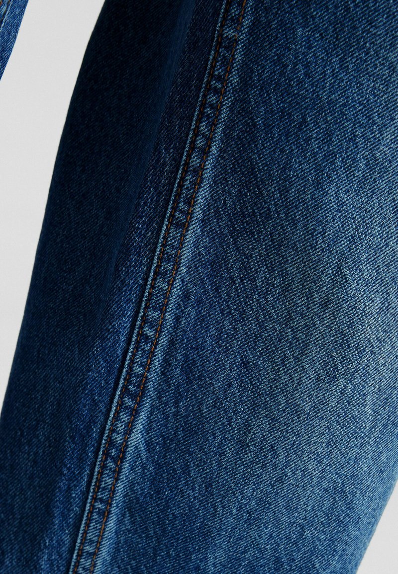 Tela de mezclilla azul oscuro con textura visible y costuras. Presenta una costura recta con hilo marrón, mostrando un primer plano de la prenda.