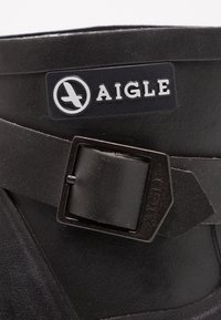 Botte en caoutchouc noire avec une finition texturée, dotée d'un logo rectangulaire Aigle et d'un détail de sangle à boucle métallique.