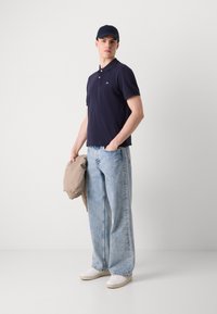 Marinblå polo skjorta med kort ärm, ljusblå byxor med vida ben, vita sneakers och en mörk keps, håller en beige jacka. Minimalistisk design.
