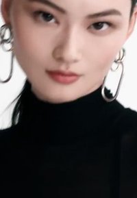 Top nero a collo alto con texture trasparente, abbinato a grandi orecchini a cerchio in argento collegati. Il modello ha capelli scuri e lisci e un trucco neutro.