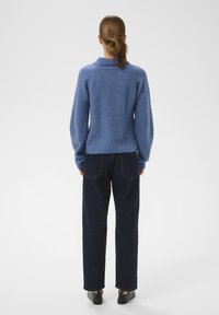 Modra pletena jopica z ohlapnim fitom, ki ima rebrasto teksturo in ovratnik. Nosita jo z temnimi kavbojkami in črnimi čevlji.