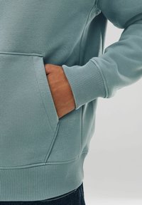 Mano parzialmente in tasca di una felpa con polsini a coste di colore azzurro chiaro, vita visibile, abbinata a jeans scuri, sfondo neutro.