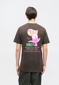 Brun bomulds-T-shirt med et farverigt grafisk motiv af en ugle på ryggen, med tekst i levende pink og grøn, der beskriver forskellige byer.
