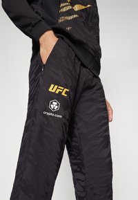 Melni sporta bikses ar tekstūru, tonālu svītru rakstu. Ar UFC logo zeltā un balto logo no crypto.com. Ar rāvējslēdzēja kabatu rokām.