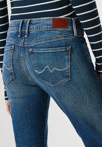 Sinine teksapükste lähipilt, mille seljataskutel on õmmeldud laineline muster, kantud tumesinist ja valget triibulist pika varrukaga särki.