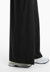 Schwarze weit geschnittene Hosen aus glattem Stoff, kombiniert mit weißen Sportschuhen, die eine strukturierte Sohle und ein atmungsaktives Mesh-Obermaterial haben.
