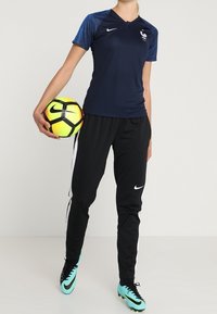 Jugadora de fútbol femenina con uniforme Nike en tonos navy y negro, sosteniendo un balón de fútbol Nike amarillo y negro, con botas turquesa, de pie frente a un fondo blanco.