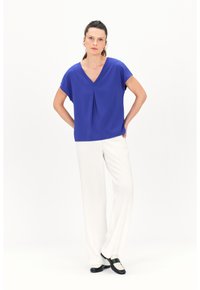 Blauwe blouse met korte mouwen en een V-hals, afgewerkt met plooidetails, gecombineerd met witte wijde broek en zwarte schoenen met witte accenten.