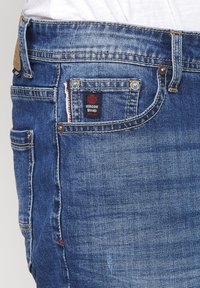 Jeans de mezclilla en lavado azul medio con un logotipo de parche oscuro, costuras en los bolsillos frontales y hilo de acento rojo. Presenta un botón de cobre y remaches.