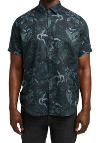 Homme portant une chemise bleu foncé à manches courtes avec un motif discret de feuilles tropicales bleu clair, associée à un pantalon noir.