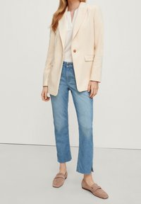 Femme portant un blazer crème sur un chemisier blanc, jean court bleu clair avec des fentes sur les côtés, et mocassins en daim beige sur fond uni.
