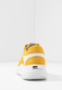 Yellow suede sneaker with white accents and thick white sole, viewed from the back on a white surface against a white corner background.
