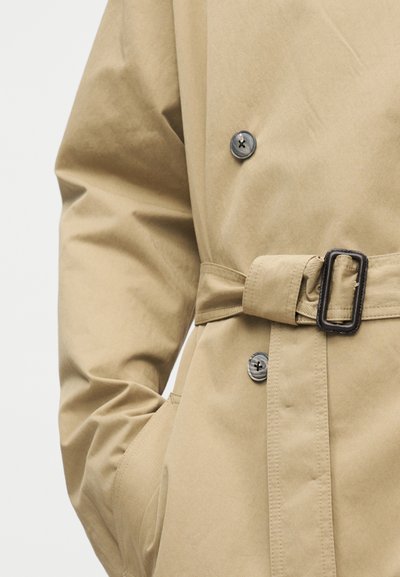 Μπεζ παλτό τύπου trench με κουμπωτό μπροστινό μέρος, ζώνη με σκοτεινή ορθογώνια αγκράφα και χέρι που είναι κρυμμένο στην πλαϊνή τσέπη.