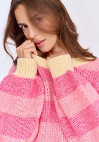 Różowy sweter w paski o luźnym kroju, z szerokimi, prążkowanymi żółtymi mankietami i dekoltem. Dzianinowa faktura z różnymi odcieniami różu.