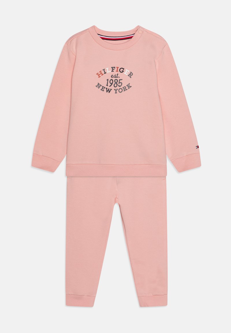 Lys rosa bomullsjakke med crew neck-genser med påtrykk av "HILFIGER est. 1985 NEW YORK" og matchende smale bukser. Ribbestrikkede mansjetter.