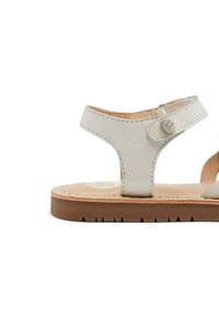 Sandal en cuir blanc avec une lanière à la cheville, fermeture à bouton circulaire et une semelle marron texturée. Présente un design plat et un style minimaliste.