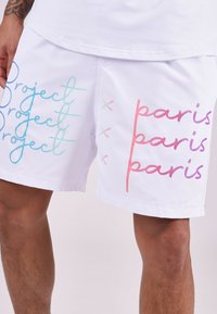 Pantalones cortos blancos con el texto impreso "Project" en azul y "paris" en rosa y morado, con acentos y diferentes estilos de fuente.