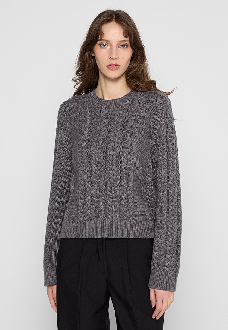 Grauer Strickpullover mit Zopfmuster, geripptem Ausschnitt und Bündchen, cropped Saum und strukturiertem Finish. Kombiniert mit schwarzen Hosen.