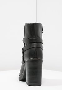 Botín de cuero negro hasta el tobillo con un tacón alto y grueso, cremallera lateral y dos correas ajustables con un detalle de logo circular.
