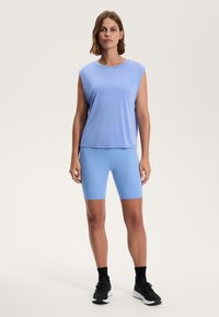 Světle modré sportovní tílko s krátkými rukávy, spárované s odpovídajícími modrými cyklistickými šortkami. Obě vyrobené z hladkého, elastického materiálu. Černé tenisky.