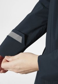 Rukáv námořnické technické bundy má reflexní šedý akcent, hladkou látku a detail s logem. Ukázaný střih je přizpůsobený a vyobrazuje držení ruky.