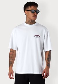 T-shirt en coton blanc à manches courtes. Présente un petit logo brodé rouge et bleu sur la poitrine. Texture lisse, coupe décontractée.