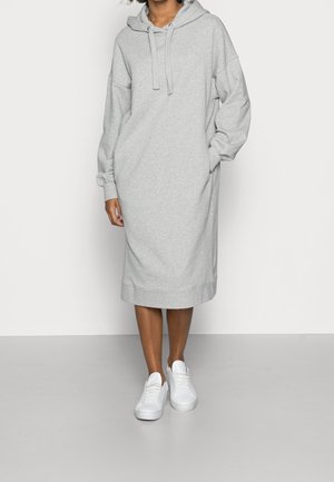 Donna che indossa un vestito con cappuccio grigio chiaro, maniche lunghe e sneakers bianche, in piedi davanti a uno sfondo bianco.